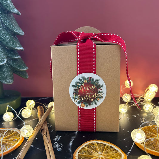 Gift box  with soap inside with a red ribbon and 'Merry Christmas' label, surrounded by festive decorations on a dark surface.