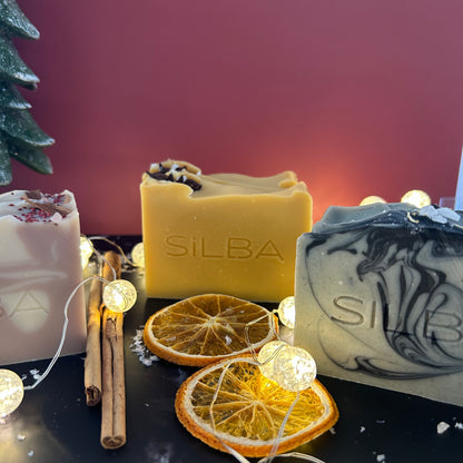 Three soaps with 'SILBA' branding on a dark surface with decorative elements like orange slices and cinnamon sticks.